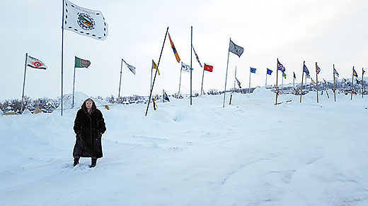 Eliida Lakota stands before flags of more than 90 Native Nations flying in defiance of the Dakota Access Pipeline route across Indian lands and under the Missouri River. Lakota traveled to Standing Rock Sioux Reservation in North Dakota to pray with the protesters, to learn and to assist with operation of the encampment. Despite a wind chill of 45 below zero, about 500 campers remain, including veterans from around the country.